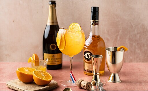 WKava cocktail: een oranje cocktail in een glas met sinaasappelschijfjes erin. Ernaast staat een fles LIcor 43, cava, een barmaatje en wat sinaasappels